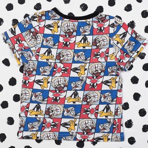 May include: A white t-shirt with a colorful cartoon print of Bugs Bunny and Daffy Duck. The print features a checkered pattern with red, blue, and yellow squares.