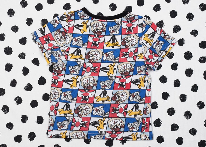 May include: A white t-shirt with a colorful cartoon print of Bugs Bunny and Daffy Duck. The print features a checkered pattern with red, blue, and yellow squares.