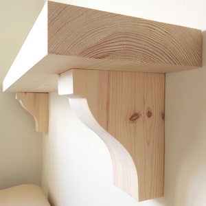 Puede incluir: Una estantería de madera con dos soportes decorativos. La estantería está hecha de madera natural y tiene un color marrón claro. Los soportes también están hechos de madera y tienen un diseño curvo.