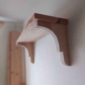 May include: A wooden shelf with decorative, curved brackets. The shelf is made of natural wood and has a light brown colour.
