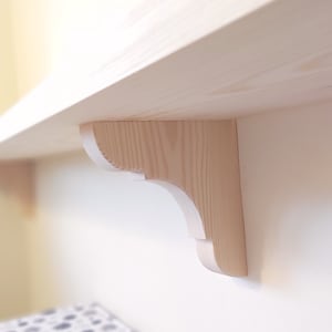 Peut inclure: Un support d'étagère en bois clair avec un design incurvé décoratif. Le support est fixé à un mur et soutient une étagère.