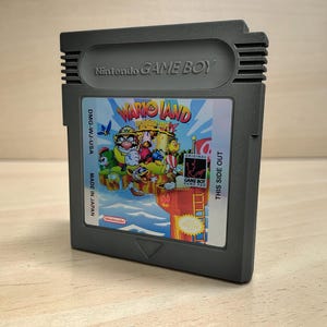 Può includere: Una cartuccia grigia per Nintendo Game Boy con il gioco "Wario Land". L'etichetta mostra personaggi dei cartoni animati colorati e il titolo del gioco. La cartuccia ha la scritta "Nintendo GAME BOY" e "MADE IN JAPAN."