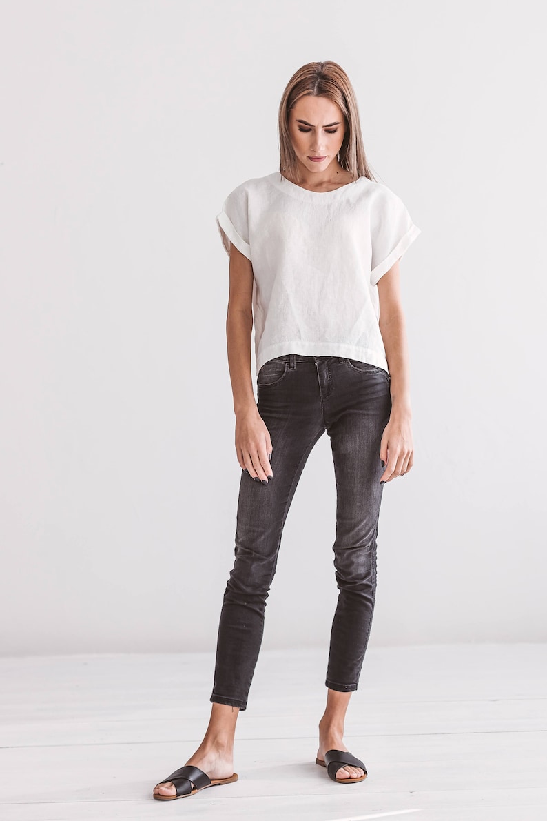 A woman wearing a white linen top with rolled sleeves and dark gray skinny jeans. The outfit is completed with black leather sandals. The background is a plain white wall and white floorboards.