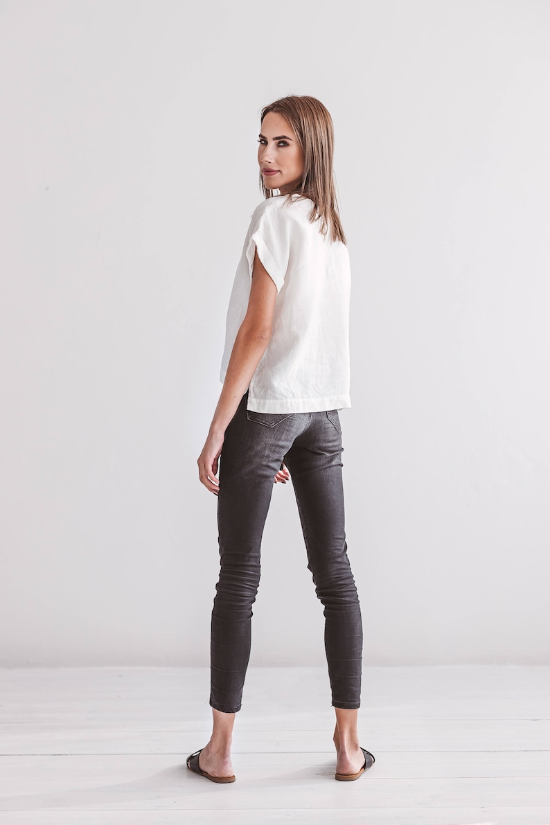 A white linen top with short sleeves and dark gray skinny jeans. The model is wearing black sandals. The outfit is simple and elegant, suitable for casual wear.