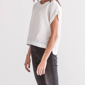 A white linen top with short sleeves and a crew neck is paired with dark gray jeans. The top has a relaxed fit and a slightly cropped length. The jeans are fitted and have a classic five-pocket design. The outfit is simple and modern.