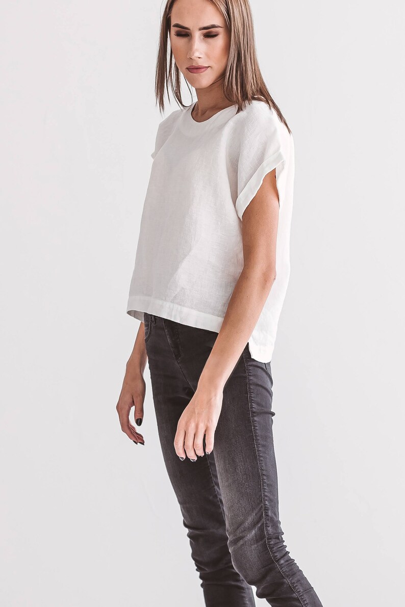 A white linen top with short sleeves and a crew neck is paired with dark gray jeans. The top has a relaxed fit and a slightly cropped length. The jeans are fitted and have a classic five-pocket design. The outfit is simple and modern.