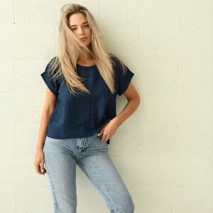 A navy blue short-sleeved top with rolled sleeves and light blue jeans. The model is leaning against a white brick wall. The top is a solid color, and the jeans are a light wash.