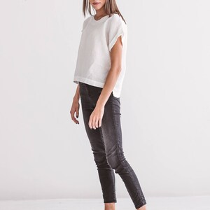 A woman wearing a white linen top with short sleeves and black jeans. The top has a round neckline and a slightly cropped fit. The jeans are a dark wash, fitted style. Black leather sandals complete the outfit. The background is a plain white wall.