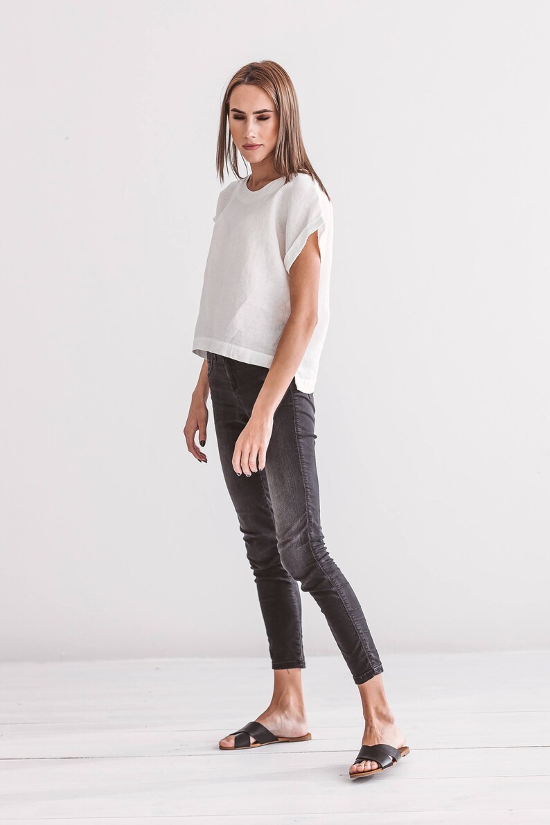 A woman wearing a white linen top with short sleeves and black jeans. The top has a round neckline and a slightly cropped fit. The jeans are a dark wash, fitted style. Black leather sandals complete the outfit. The background is a plain white wall.