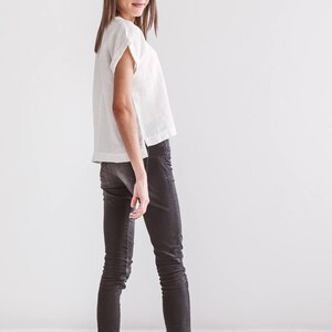 A woman wearing a white linen top with short sleeves and dark gray skinny jeans. The outfit is paired with black sandals. The background is a plain white wall.