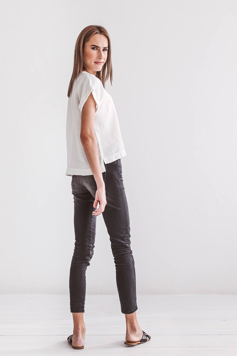 A woman wearing a white linen top with short sleeves and dark gray skinny jeans. The outfit is paired with black sandals. The background is a plain white wall.