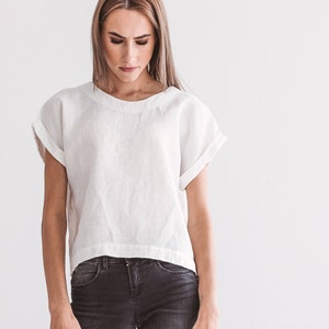 A white linen top with rolled sleeves and a rounded neckline. The top is paired with dark gray denim jeans. The model is looking down. The background is a plain white wall. The overall style is minimalist and casual.