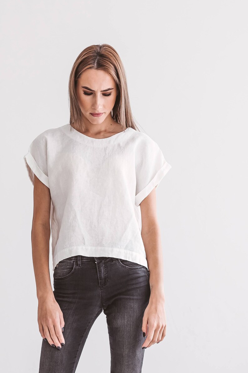 A white linen top with rolled sleeves and a rounded neckline. The top is paired with dark gray denim jeans. The model is looking down. The background is a plain white wall. The overall style is minimalist and casual.