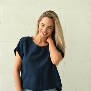 A navy blue, short-sleeved top with a round neckline. The top is paired with light blue denim jeans. The model is smiling and standing in front of a white brick wall. The top appears to be made of linen.
