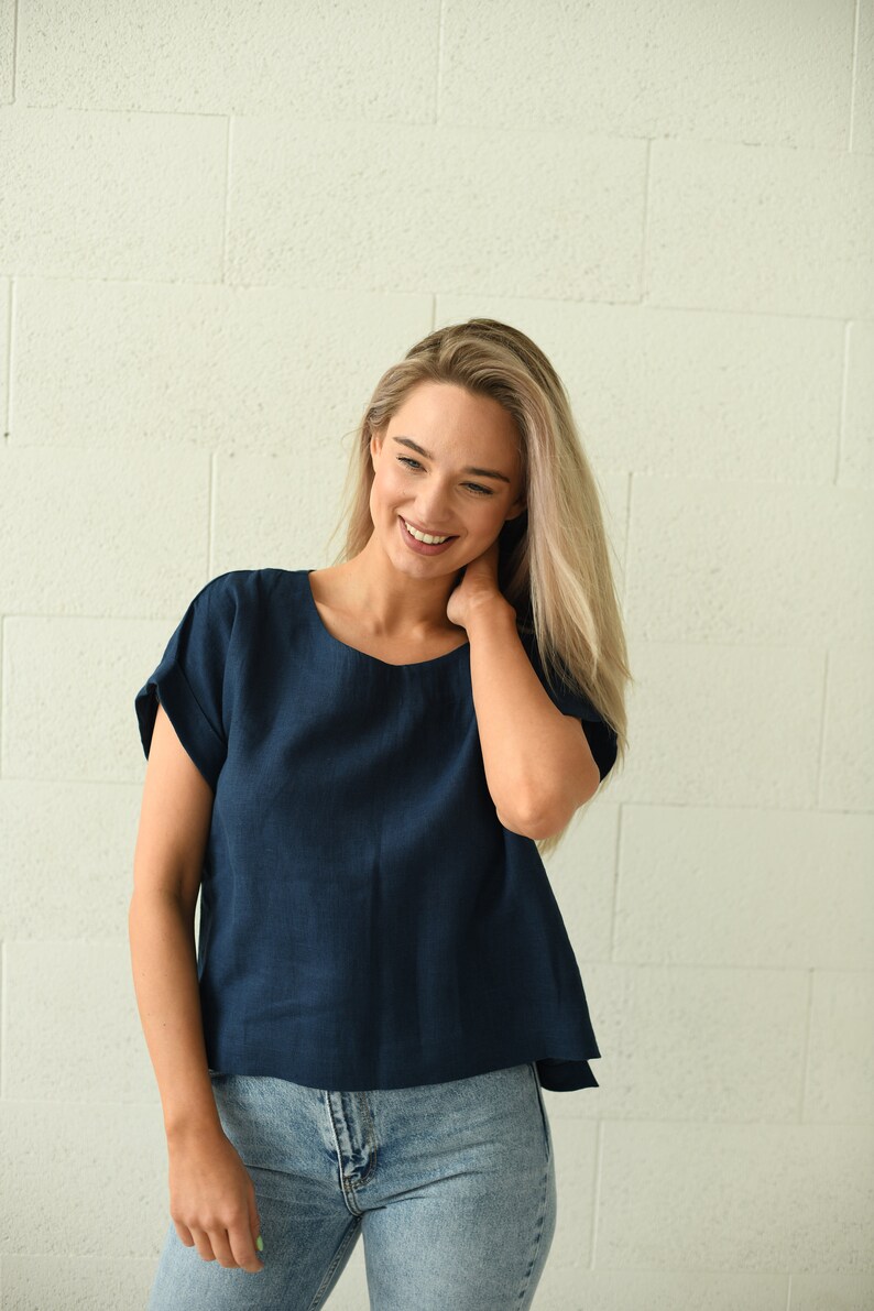 A navy blue, short-sleeved top with a round neckline. The top is paired with light blue denim jeans. The model is smiling and standing in front of a white brick wall. The top appears to be made of linen.