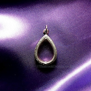 Op de afbeelding: Een zilverkleurige hanger in de vorm van een druppel, met een sierlijke, gegraveerde rand. De hanger hangt aan een kleine lus en ligt op een paarse satijnen stof. Het ontwerp is elegant en gedetailleerd.