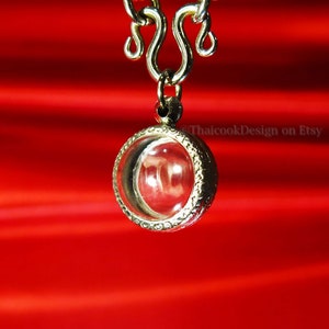 May include: A silver chain necklace with a round pendant featuring a clear glass insert. The pendant has a decorative border and is hanging against a red background.