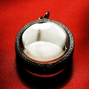 Puede incluir: Un medallón de plata con un inserto redondo de vidrio transparente y un borde decorativo. El medallón está sobre un fondo rojo.