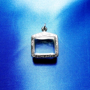 Puede incluir: Un medallón de plata con un diseño adornado y un inserto de vidrio transparente. El medallón está colgado sobre un fondo azul.