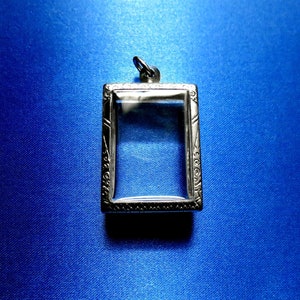 Może przedstawiać: Srebrny wisiorek z przezroczystym wstawką ze szkła. Wisiorek ma ozdobną ramkę i pętelkę u góry do przymocowania łańcuszka.