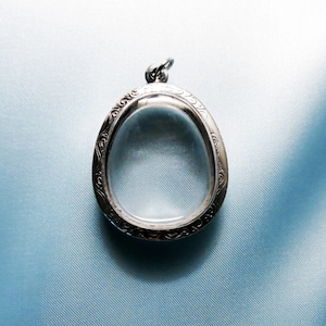 Op de afbeelding: Een zilveren ovale hanger met een versierd ontwerp en een transparante glazen inzet.