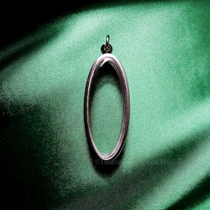 Op de afbeelding: Een zilveren ovale locket-hanger met een glad, reflecterend oppervlak. De hanger hangt tegen een groene stoffen achtergrond.