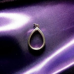 Puede incluir: Un colgante de plata en forma de lágrima con un inserto de vidrio transparente. El colgante está sobre un fondo de satén morado.