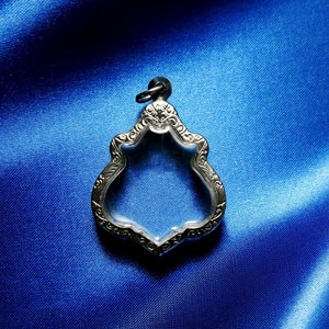 Puede incluir: Un colgante de plata con un diseño adornado y un centro de vidrio transparente. El colgante está sobre un fondo de satén azul.