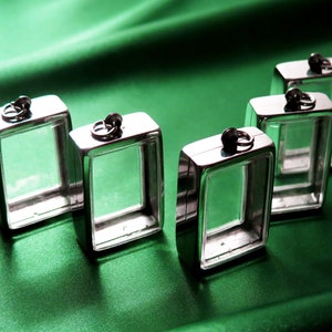 May include: Six silver-toned square pendant charms with clear glass inserts. The charms are arranged in a row on a green fabric surface.