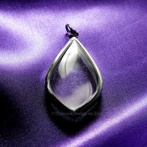 Puede incluir: Un colgante de plata en forma de lágrima con un inserto de vidrio transparente. El colgante está sobre un fondo de satén morado.