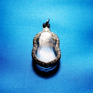 Puede incluir: Un colgante de plata con un inserto de vidrio transparente, que presenta un diseño intrincado alrededor del borde. El colgante está sobre un fondo azul.