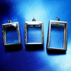 May include: Three silver-toned pendant frames with clear glass inserts. The frames have ornate designs and are arranged in a row on a blue background.