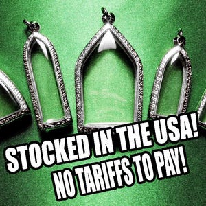 May include: Several silver-colored, arched pendants with clear centers are displayed on a green background. The pendants have ornate, decorative borders. The image includes the text "STOCKED IN THE USA! NO TARIFFS TO PAY!"