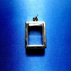 May include: A rectangular silver pendant with an ornate border and a clear centre, suspended from a small loop. The pendant is set against a vibrant blue background, creating a striking contrast.