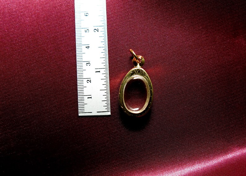 K&ouml;nnte beinhalten: Ein ovaler Anh&auml;nger aus Gold mit einem transparenten Glaseinsatz. Der Anh&auml;nger ist etwa 3,8 cm lang und hat ein filigranes Design.