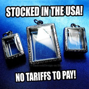 May include: Three silver-toned rectangular pendants with clear glass inserts. The pendants have ornate, decorative frames and small loops at the top for hanging. The text "STOCKED IN THE USA!" and "NO TARIFFS TO PAY!" are displayed in white.