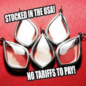 May include: Five teardrop-shaped pendants with silver-colored frames and clear glass inserts. The pendants are arranged on a red background. Text in the image reads "STOCKED IN THE USA!" and "NO TARIFFS TO PAY!"