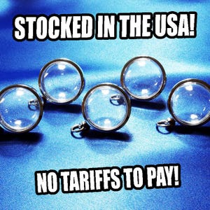 May include: Five clear, round glass pendants with silver-colored metal rims and small loops, arranged on a blue satin fabric. The text "STOCKED IN THE USA!" and "NO TARIFFS TO PAY!" are displayed in white block letters.