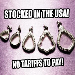 May include: Five silver-toned teardrop-shaped pendants with clear centers are displayed on a textured purple surface. The pendants vary in size and design, with some featuring decorative embellishments. Text at the top reads "STOCKED IN THE USA!" and at the bottom "NO TARIFFS TO PAY!"