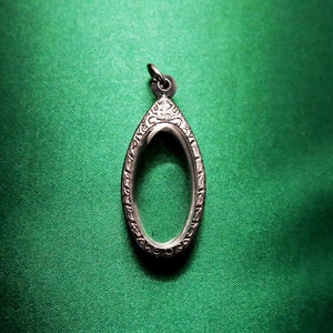 May include: Silver pendant in an elongated oval shape. The pendant has an open centre and a decorative, patterned border. It has a small loop at the top for a chain. The pendant is set against a green background.