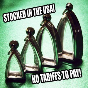 May include: Four clear, arched pendants with silver-colored frames and black tops are arranged on a green surface. The text "STOCKED IN THE USA!" and "NO TARIFFS TO PAY!" are displayed in white.