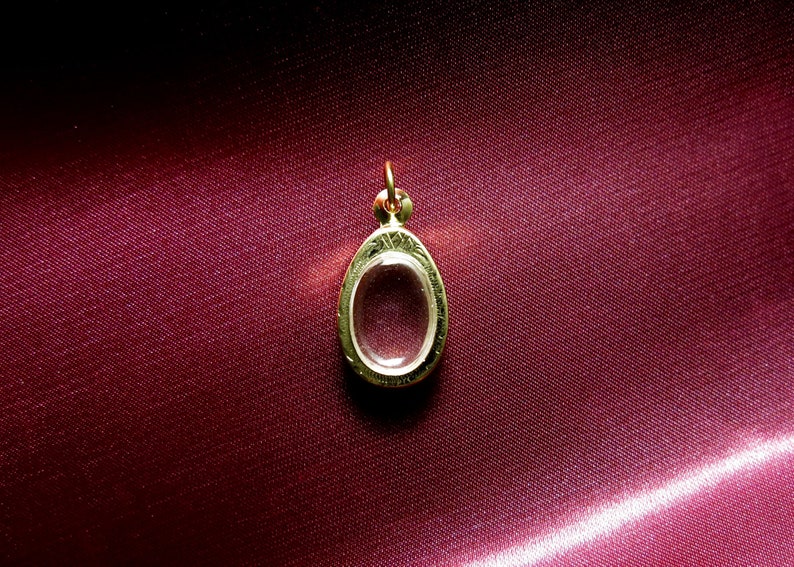 K&ouml;nnte beinhalten: Ein goldener Anh&auml;nger mit einem klaren, ovalen Glaseinsatz. Der Anh&auml;nger liegt auf einem roten Stoffhintergrund.