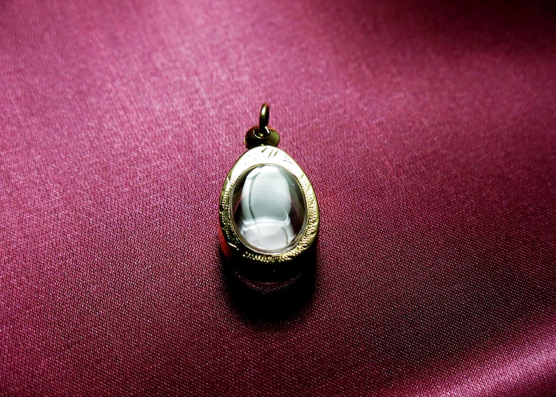 K&ouml;nnte beinhalten: Ein ovaler Anh&auml;nger aus Gold mit einem transparenten Glaseinsatz. Der Anh&auml;nger liegt auf einem burgunderfarbenen Stoffhintergrund.