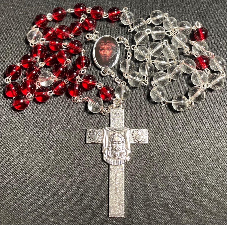 May include: A rosary with red and clear beads. The rosary features a silver cross pendant with a detailed image of Jesus and a small oval pendant with a portrait of Jesus. The beads are strung on a silver chain.
