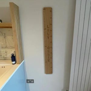 Peut inclure: Une toise en bois verticale avec des graduations grav&eacute;es et les pr&eacute;noms LILY et OLLIE inscrits en lettres sombres. Cette planche en bois clair est fix&eacute;e au mur et affiche des mesures en centim&egrave;tres le long de son bord lat&eacute;ral.