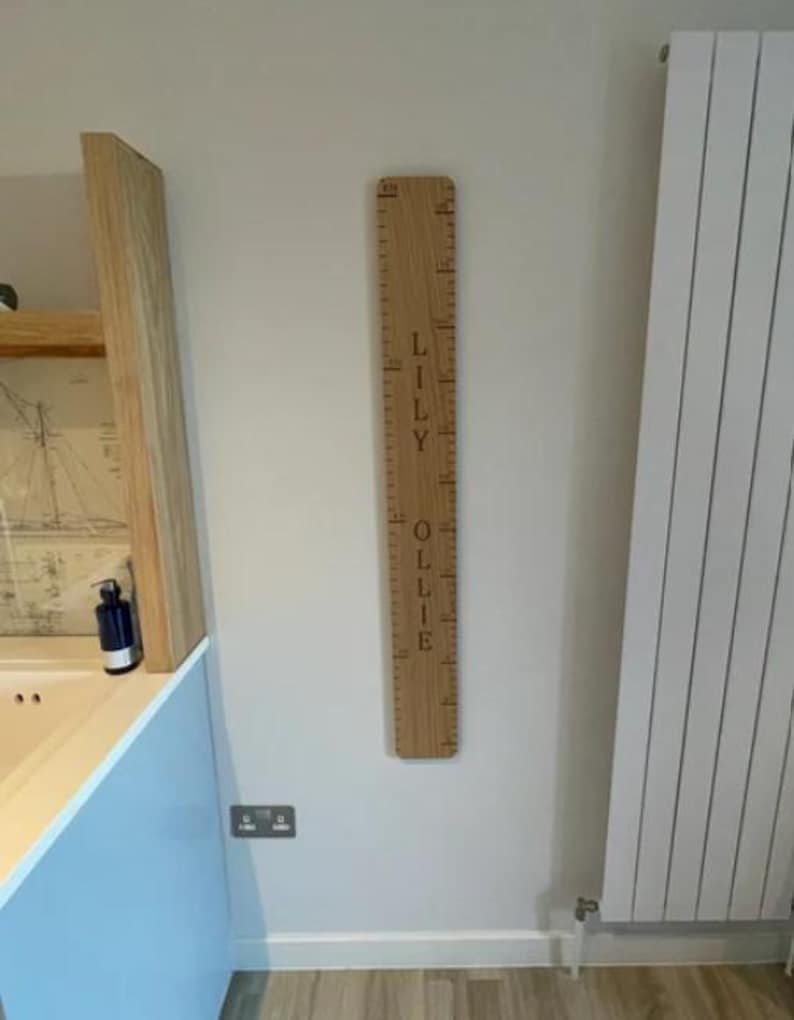Peut inclure: Une toise en bois verticale avec des graduations grav&eacute;es et les pr&eacute;noms LILY et OLLIE inscrits en lettres sombres. Cette planche en bois clair est fix&eacute;e au mur et affiche des mesures en centim&egrave;tres le long de son bord lat&eacute;ral.