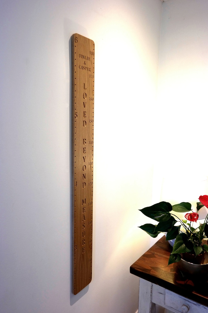 Peut inclure: Une toise murale en bois vertical avec des graduations grav&eacute;es en pieds et en centim&egrave;tres. La planche pr&eacute;sente le texte "FINLAY & CASPER" et "LOVED BEYOND MEASURE" dans une police sombre et creus&eacute;e, avec une finition en bois clair naturel.
