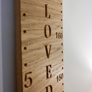 Peut inclure: Une toise en bois brun clair avec un motif de grain vertical. Le mot LOVED est grav&eacute; en grandes lettres majuscules au centre. Des marquages num&eacute;riques pour 150, 160 et 170 sont visibles &agrave; c&ocirc;t&eacute; d'une s&eacute;rie de petits trous perc&eacute;s.