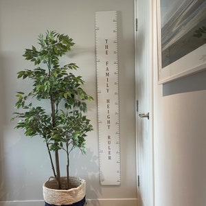 Peut inclure: Une règle de mesure de la taille familiale en bois blanc avec du texte marron et des marquages de mesure en centimètres. La règle est fixée au mur. Une plante verte à feuilles dans un panier tressé est à côté de la règle. Le texte dit "THE FAMILY HEIGHT RULER".