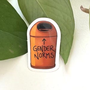 Könnte beinhalten: Ein orangefarbener Aufkleber mit einer Cartoon-Mülltonne mit den Worten "GENDER NORMS" und einem Pfeil nach oben. Der Aufkleber befindet sich vor einem weißen Hintergrund mit grünen Pflanzenblättern.